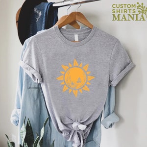 Puede incluir: Una camiseta gris con un gr&aacute;fico de sol amarillo. El sol tiene una cara y est&aacute; rodeado de rayos. La camiseta est&aacute; en una percha.
