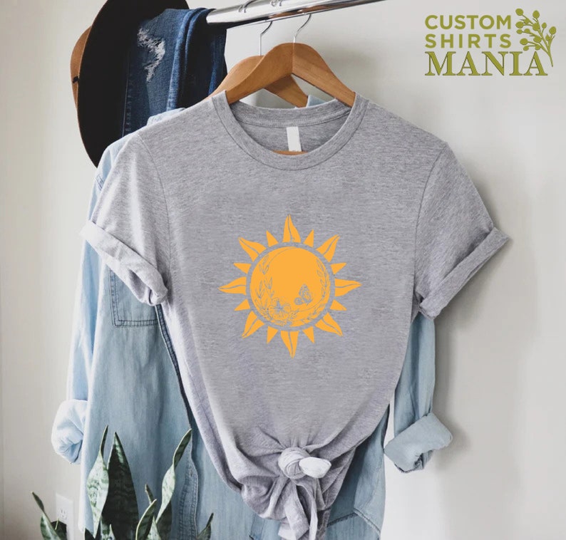 Puede incluir: Una camiseta gris con un gr&aacute;fico de sol amarillo. El sol tiene una cara y est&aacute; rodeado de rayos. La camiseta est&aacute; en una percha.