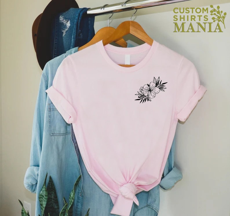 May include: A pink t-shirt with a black floral design on the left chest. The shirt is hanging on a hanger with a denim shirt and a hat behind it.