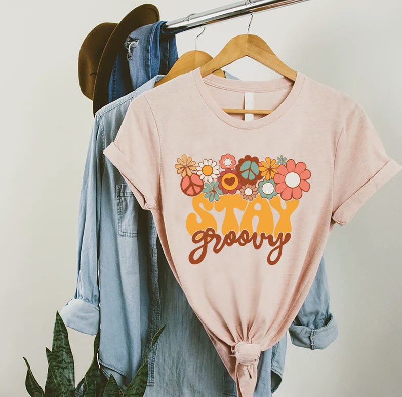 May include: A pink t-shirt with a retro-style graphic design featuring flowers, peace signs, and the text "Stay groovy".