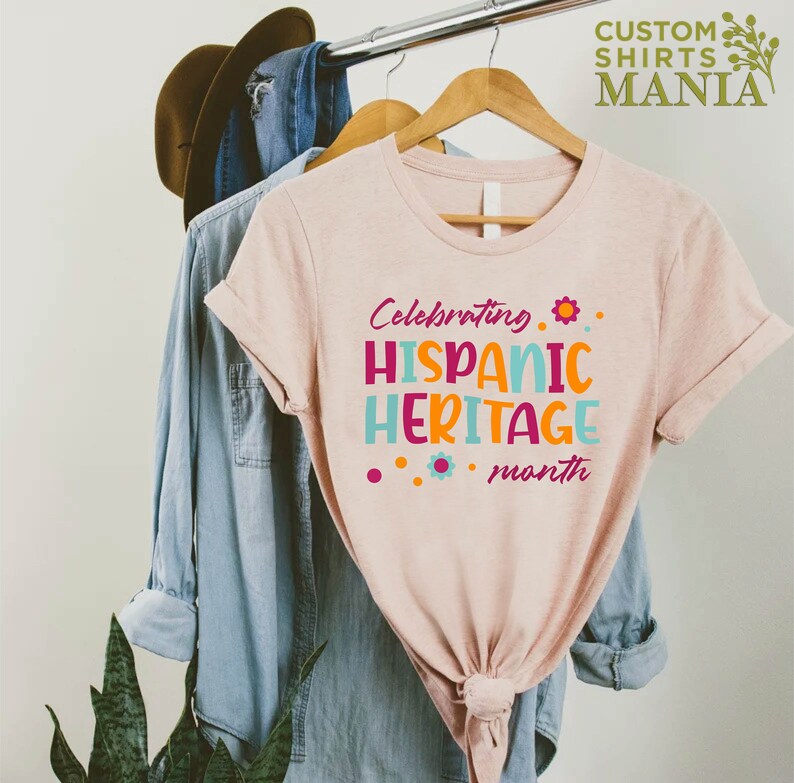 May include: A pink t-shirt with the text "Celebrating Hispanic Heritage month" in colorful letters and a small flower graphic.