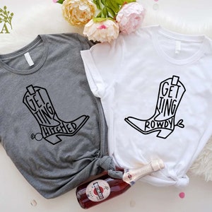 May include: Two t-shirts with a black boot graphic and text. The grey shirt says "Getting Hitched" and the white shirt says "Getting Rowdy".
