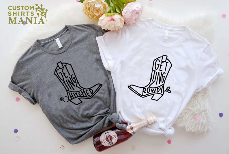 May include: Two t-shirts with a black boot graphic and text. The grey shirt says "Getting Hitched" and the white shirt says "Getting Rowdy".