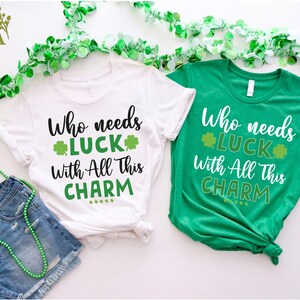 May include: Two white t-shirts with green shamrock designs. The text on the shirts reads "Who needs luck with all this charm".