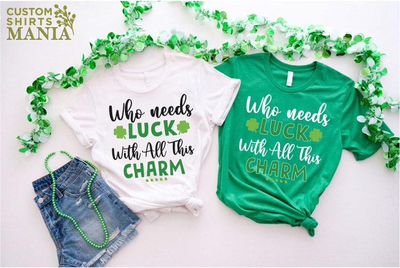 May include: Two white t-shirts with green shamrock designs. The text on the shirts reads "Who needs luck with all this charm".