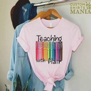 May include: Pink t-shirt with the text "Teaching with flair" and a row of colorful markers.