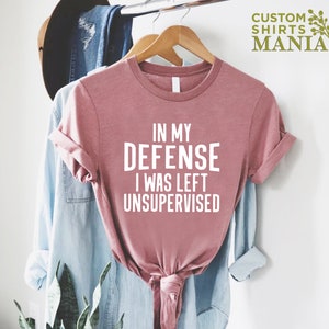 May include: A pink t-shirt with the text "In my defense I was left unsupervised" printed in white.