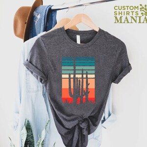 May include: A dark gray t-shirt with a retro-style graphic of a cactus silhouette against a sunset-colored striped background. The text "CUSTOM SHIRTS MANIA" is visible in the top right corner of the image.