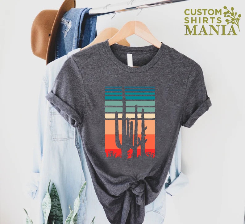 May include: A dark gray t-shirt with a retro-style graphic of a cactus silhouette against a sunset-colored striped background. The text "CUSTOM SHIRTS MANIA" is visible in the top right corner of the image.