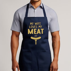 May include: A navy blue apron with a large pocket on the front. The apron has the text "My wife loves my meat" in gold lettering with a graphic of a hot dog and fork.
