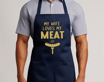 May include: A navy blue apron with a large pocket on the front. The apron has the text "My wife loves my meat" in gold lettering with a graphic of a hot dog and fork.