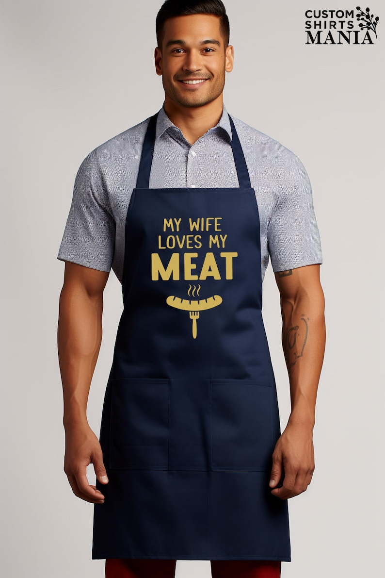 May include: A navy blue apron with a large pocket on the front. The apron has the text "My wife loves my meat" in gold lettering with a graphic of a hot dog and fork.