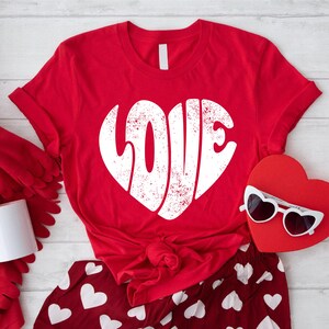 May include: Red t-shirt with a white heart-shaped graphic that says "LOVE" in a retro font. The shirt is laid out on a white wooden surface with red and white heart-patterned pajamas and a red heart-shaped box with white sunglasses.