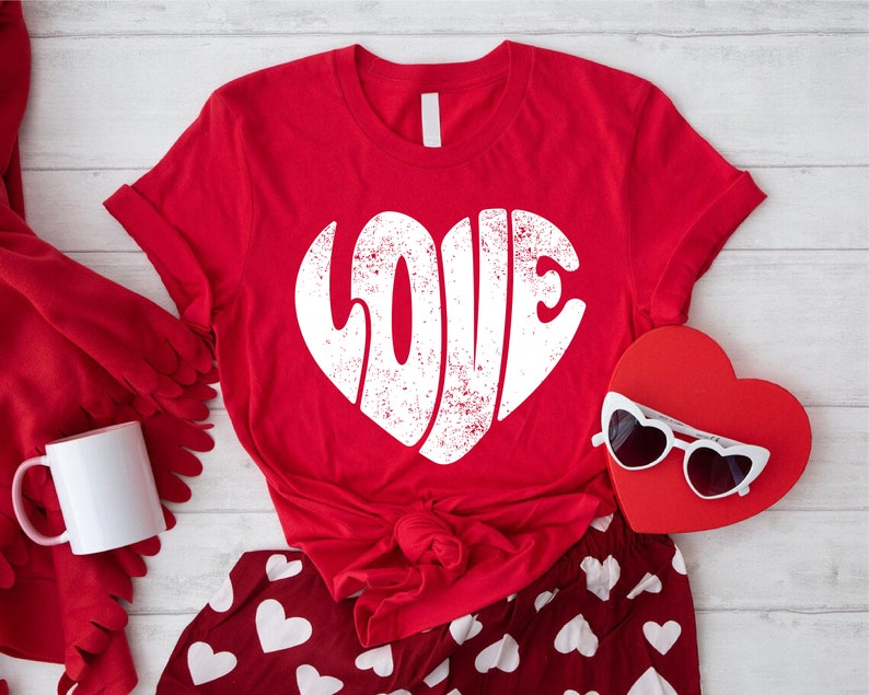 May include: Red t-shirt with a white heart-shaped graphic that says "LOVE" in a retro font. The shirt is laid out on a white wooden surface with red and white heart-patterned pajamas and a red heart-shaped box with white sunglasses.