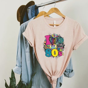May include: A pink t-shirt with a graphic design of a heart with a leopard print, a cassette tape, a disco ball, and the text "I love the 80s".