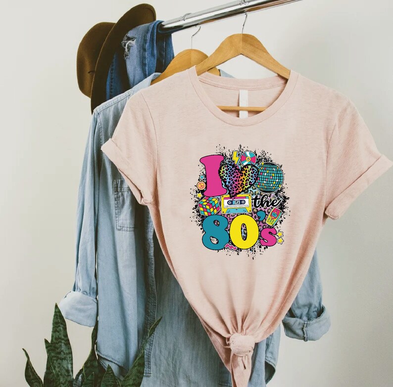 May include: A pink t-shirt with a graphic design of a heart with a leopard print, a cassette tape, a disco ball, and the text "I love the 80s".