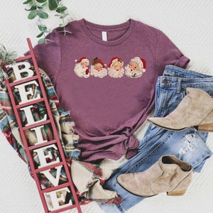 May include: A purple t-shirt with a graphic of four vintage Santa Claus faces. The t-shirt is displayed on a white surface with a wooden sign that reads "BELIEVE" and a pair of light brown ankle boots.