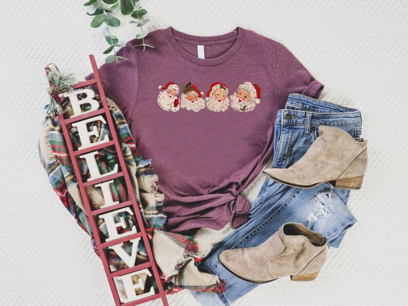 May include: A purple t-shirt with a graphic of four vintage Santa Claus faces. The t-shirt is displayed on a white surface with a wooden sign that reads "BELIEVE" and a pair of light brown ankle boots.