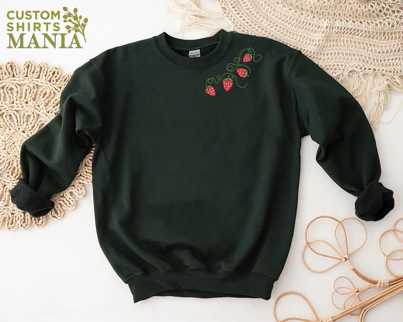 Puede incluir: Sudadera verde oscuro con fresas bordadas y el texto "CUSTOM SHIRTS MANIA" en verde. Las fresas son rojas con tallos y hojas verdes. La sudadera tiene cuello redondo y mangas largas.