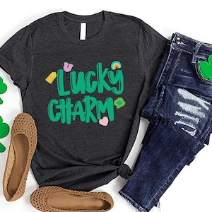 May include: A dark grey t-shirt with the text "Lucky Charm" in green lettering. The shirt has a variety of small green, yellow, pink, and rainbow coloured graphics around the text.
