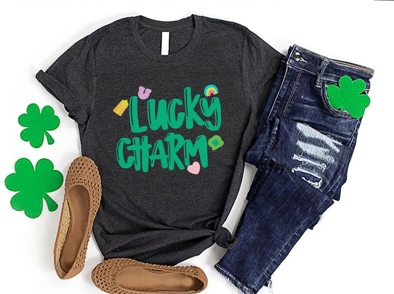 May include: A dark grey t-shirt with the text "Lucky Charm" in green lettering. The shirt has a variety of small green, yellow, pink, and rainbow coloured graphics around the text.