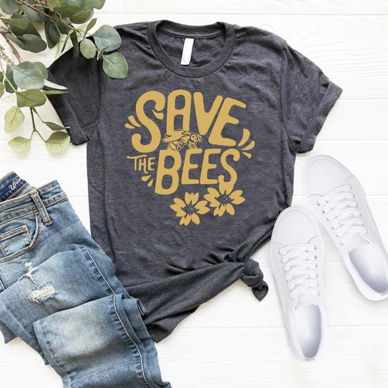 May include: Dark gray t-shirt with the text "SAVE THE BEES" in gold, featuring a bee and flowers. The shirt is paired with blue jeans and white sneakers on a white wooden surface.