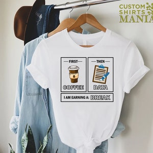 May include: White t-shirt with a graphic of a coffee cup and a clipboard with a pen. The text reads "First - Coffee, Then - Data, I am earning a: Break".