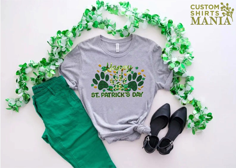 May include: A gray t-shirt with a green shamrock and paw print design. The shirt says "Happy St. Patrick's Day".
