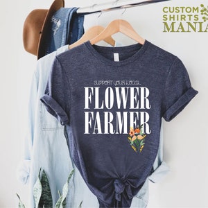 May include: A navy blue t-shirt with white text that reads "Support Your Local Flower Farmer". The text is centered on the shirt and is surrounded by a small bouquet of flowers.