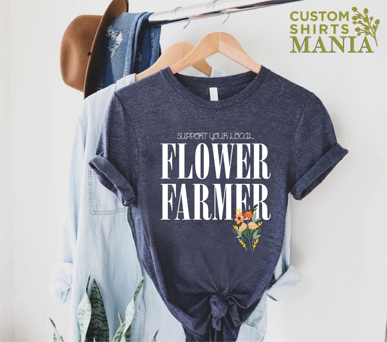 May include: A navy blue t-shirt with white text that reads "Support Your Local Flower Farmer". The text is centered on the shirt and is surrounded by a small bouquet of flowers.