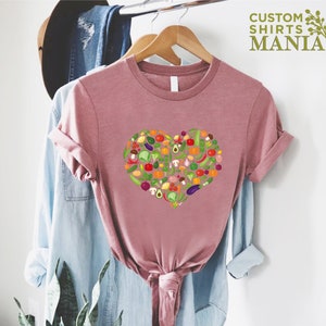 May include: Dusty rose t-shirt featuring a heart-shaped design composed of various colorful vegetables. The shirt is hanging on a wooden hanger. The text "CUSTOM SHIRTS MANIA" is printed in green on the upper right.