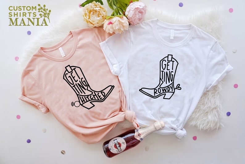 May include: Two white t-shirts with black boot designs and text. One shirt says "Getting Hitched" and the other says "Getting Rowdy".