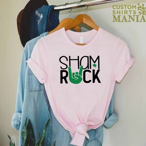 Peut inclure: T-shirt rose avec un texte noir qui dit "Shamrock" avec un geste de main rock and roll vert et un tr&egrave;fle.