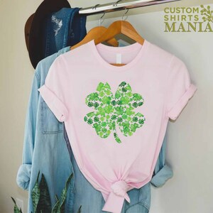 May include: A pink t-shirt with a green four-leaf clover design made of smaller clovers. The design is centered on the shirt. The shirt is hanging on a hanger.