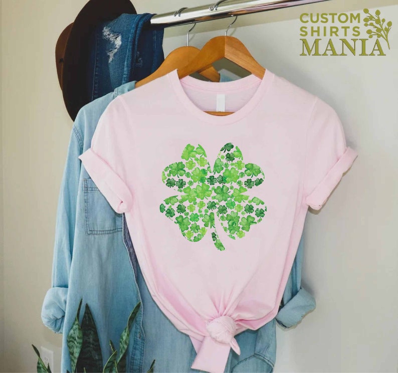 May include: A pink t-shirt with a green four-leaf clover design made of smaller clovers. The design is centered on the shirt. The shirt is hanging on a hanger.