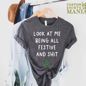 May include: A dark gray t-shirt with white text that reads "Look at me being all festive and shit" with a small green Christmas tree with lights below the text.