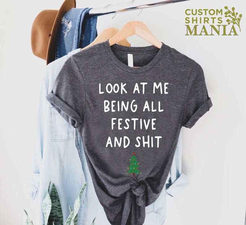 May include: A dark gray t-shirt with white text that reads "Look at me being all festive and shit" with a small green Christmas tree with lights below the text.
