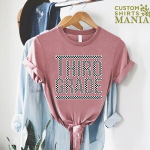 Pu&ograve; includere: Una maglietta rosa con un design a scacchi bianco e nero che dice "TERZA CLASSE" su di essa.