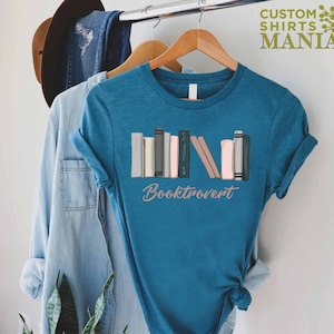 May include: A teal blue t-shirt with a graphic of a stack of colorful books and the text "Booktrovert".