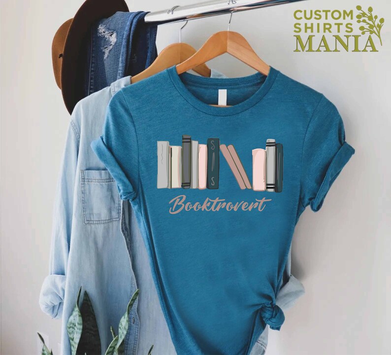 May include: A teal blue t-shirt with a graphic of a stack of colorful books and the text "Booktrovert".