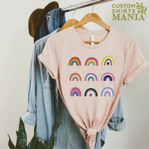 K&ouml;nnte beinhalten: Ein pinkfarbenes T-Shirt mit einem Design aus neun verschiedenen Regenbogenflaggen. Das Shirt h&auml;ngt an einem Kleiderb&uuml;gel und hat einen geknoteten Saum.