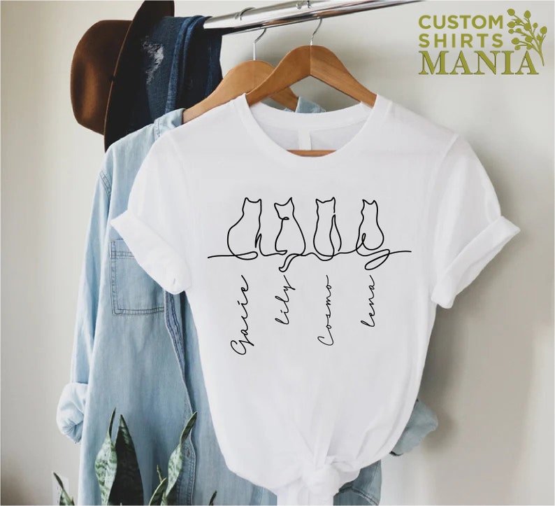 May include: White t-shirt with a line drawing of four cats and the names "Gracie", "Lily", "Cosmo", and "Lena" written underneath.