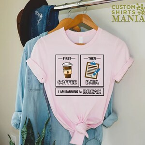 May include: Pink t-shirt with a graphic design that says "First Coffee, Then Data, I am earning a Break". The graphic features a cup of coffee and a clipboard with a pen.