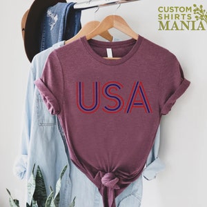 Może przedstawiać: Bordowy t-shirt z nadrukiem słowa "USA" w czerwono-niebieskie paski. Koszulka wisi na drewnianym wieszaku.