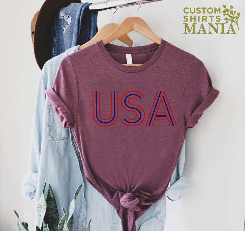 Może przedstawiać: Bordowy t-shirt z nadrukiem słowa "USA" w czerwono-niebieskie paski. Koszulka wisi na drewnianym wieszaku.