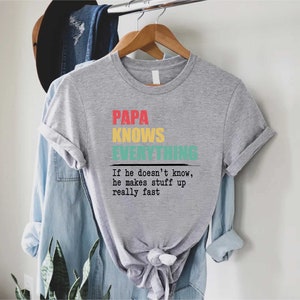 May include: A grey t-shirt with the words "PAPA KNOWS EVERYTHING" in red, yellow, and teal. Underneath is the phrase, "If he doesn't know, he makes stuff up really fast."