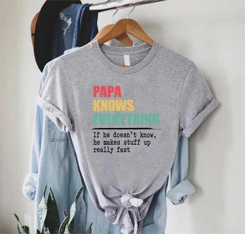 May include: A grey t-shirt with the words "PAPA KNOWS EVERYTHING" in red, yellow, and teal. Underneath is the phrase, "If he doesn't know, he makes stuff up really fast."