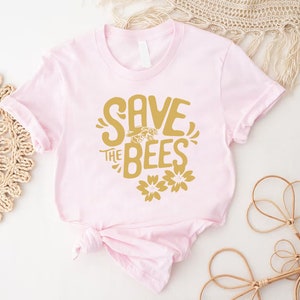 May include: A light pink t-shirt with a gold graphic that reads "SAVE THE BEES" with a bee and flower illustrations. The shirt is knotted at the bottom and is displayed on a white surface with decorative items.