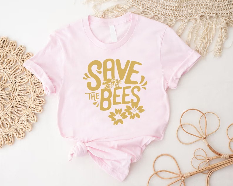 May include: A light pink t-shirt with a gold graphic that reads "SAVE THE BEES" with a bee and flower illustrations. The shirt is knotted at the bottom and is displayed on a white surface with decorative items.