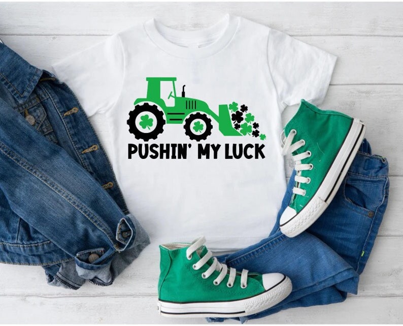 May include: A white t-shirt with a green tractor and shamrocks printed on it. The text "Pushin' My Luck" is printed below the tractor. The shirt is paired with blue jeans and green sneakers.